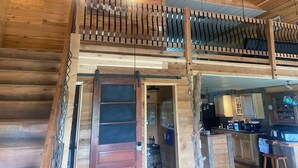 A wooden cabin interior with a loft bedroom, a kitchen area, and a dining space.