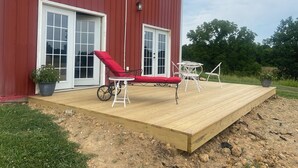 A red barn with a wooden deck, a red lounge chair, a white table and chairs, and a potted plant.