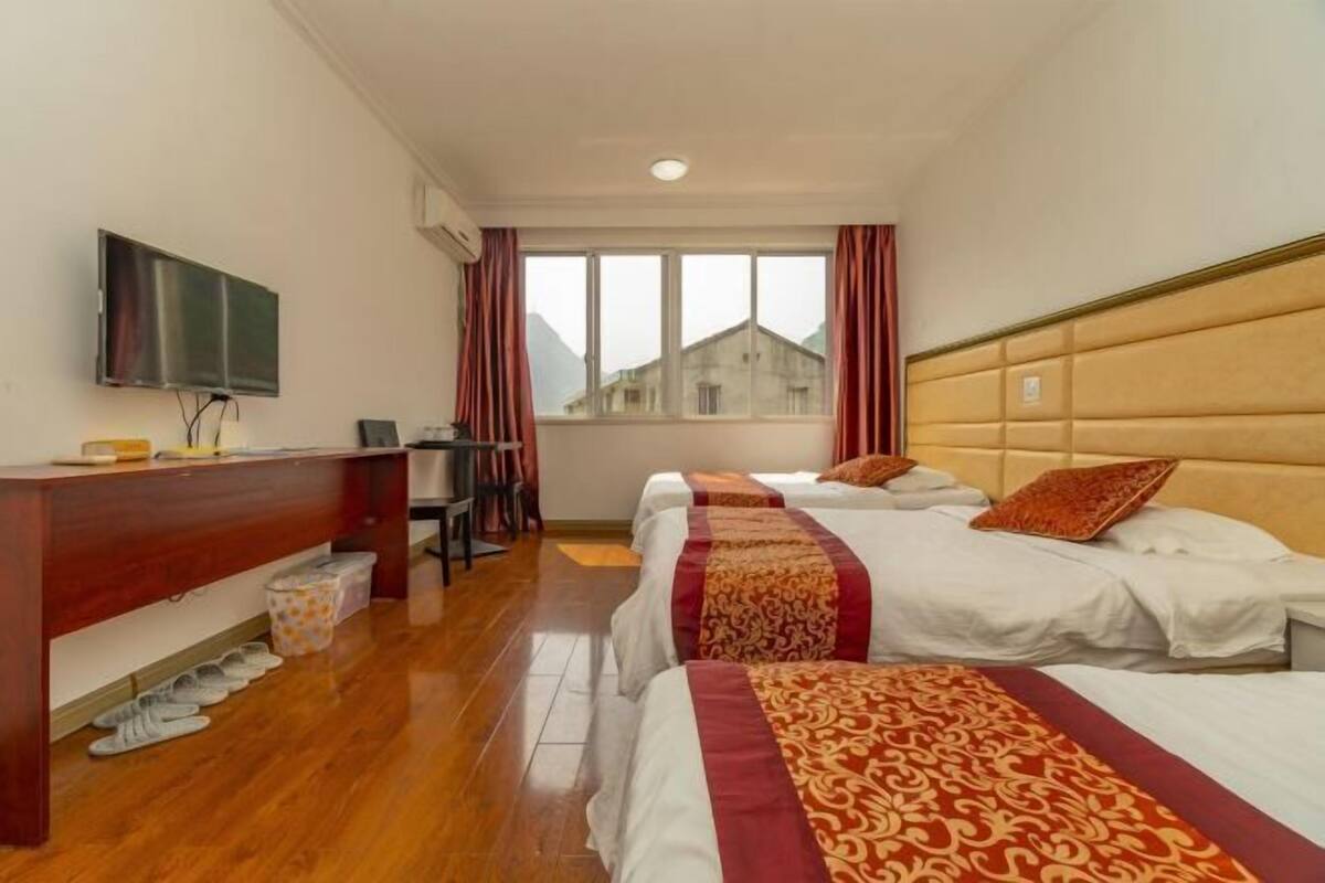 A hotel room with a wooden desk, a flat-screen TV, two beds with patterned bedspreads, a window with curtains, and a view of a building outside.
