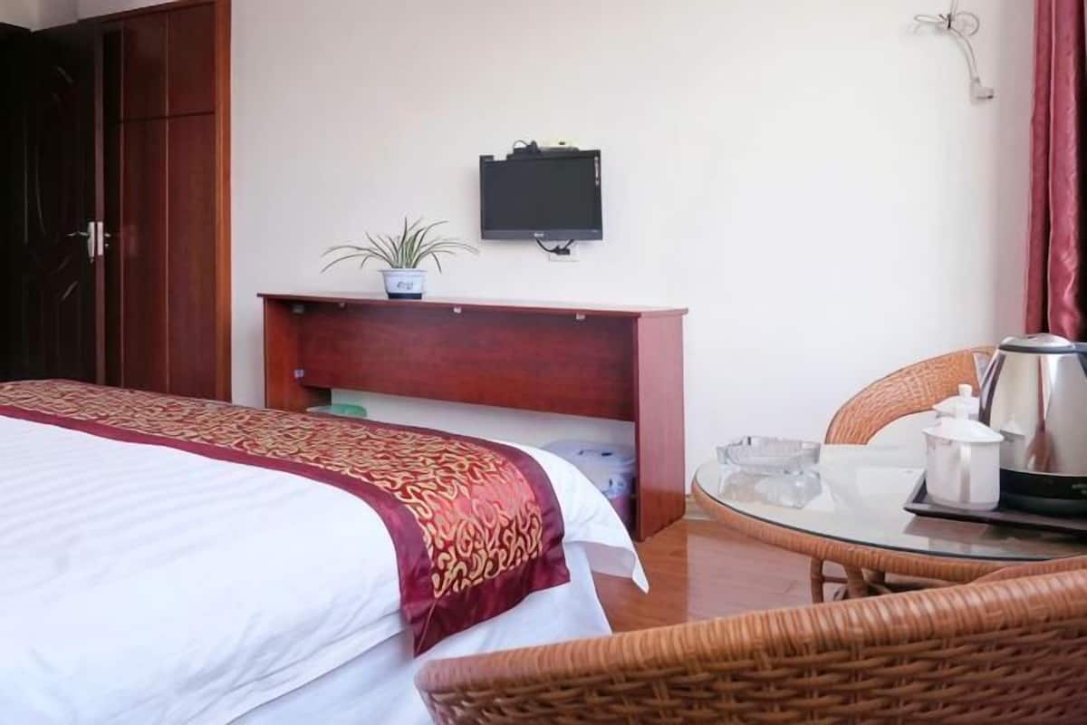 A hotel room with a bed, a small table with a kettle, a chair, a TV on the wall, and a potted plant.
