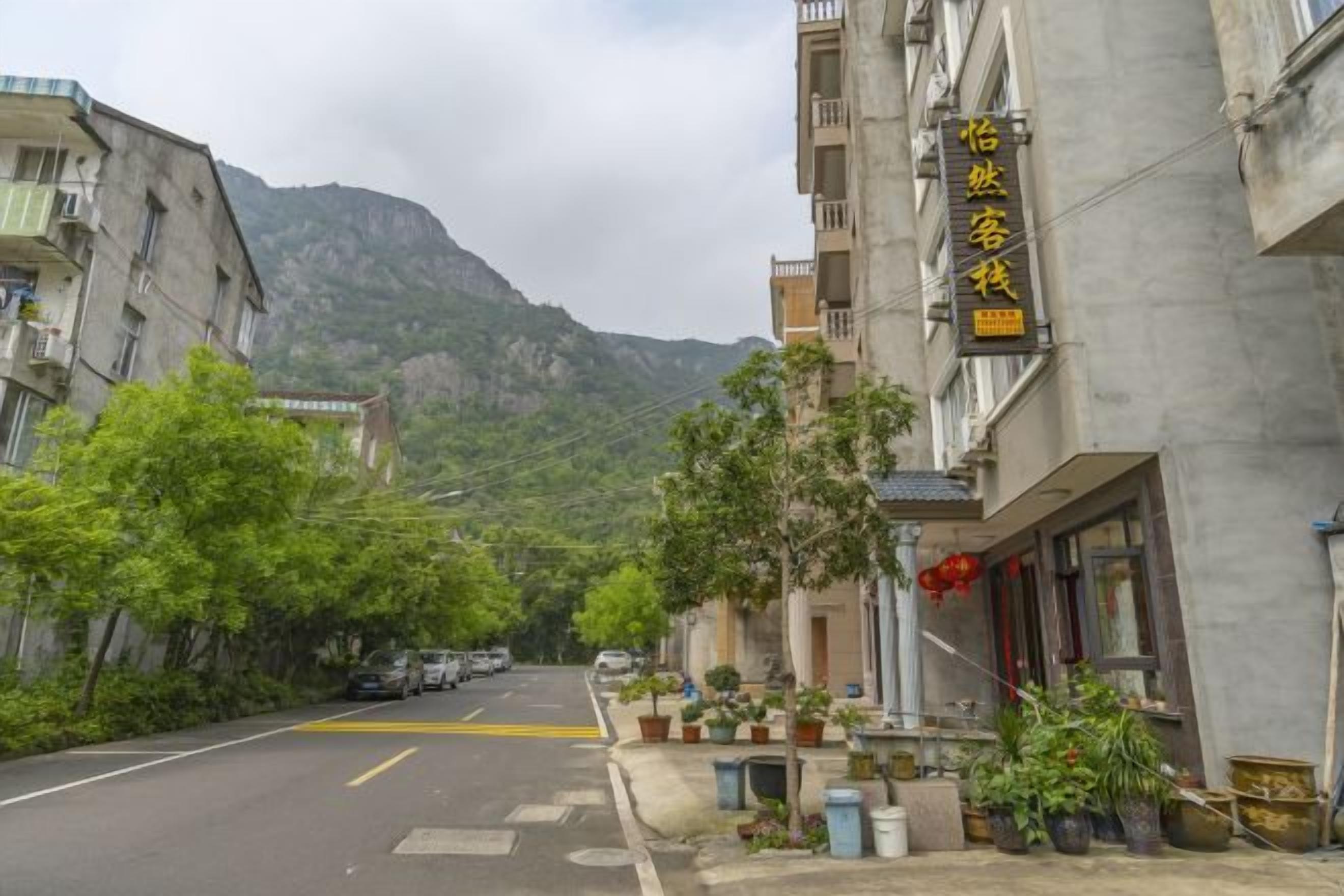 A street view with a building featuring a sign in a foreign language, a mountain in the background, and parked cars along the road.
