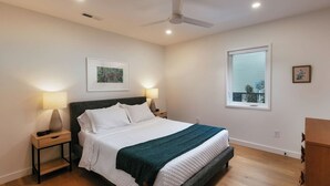 Un dormitorio con cama, lámparas de noche, ventilador de techo, ventana y un cuadro en la pared.