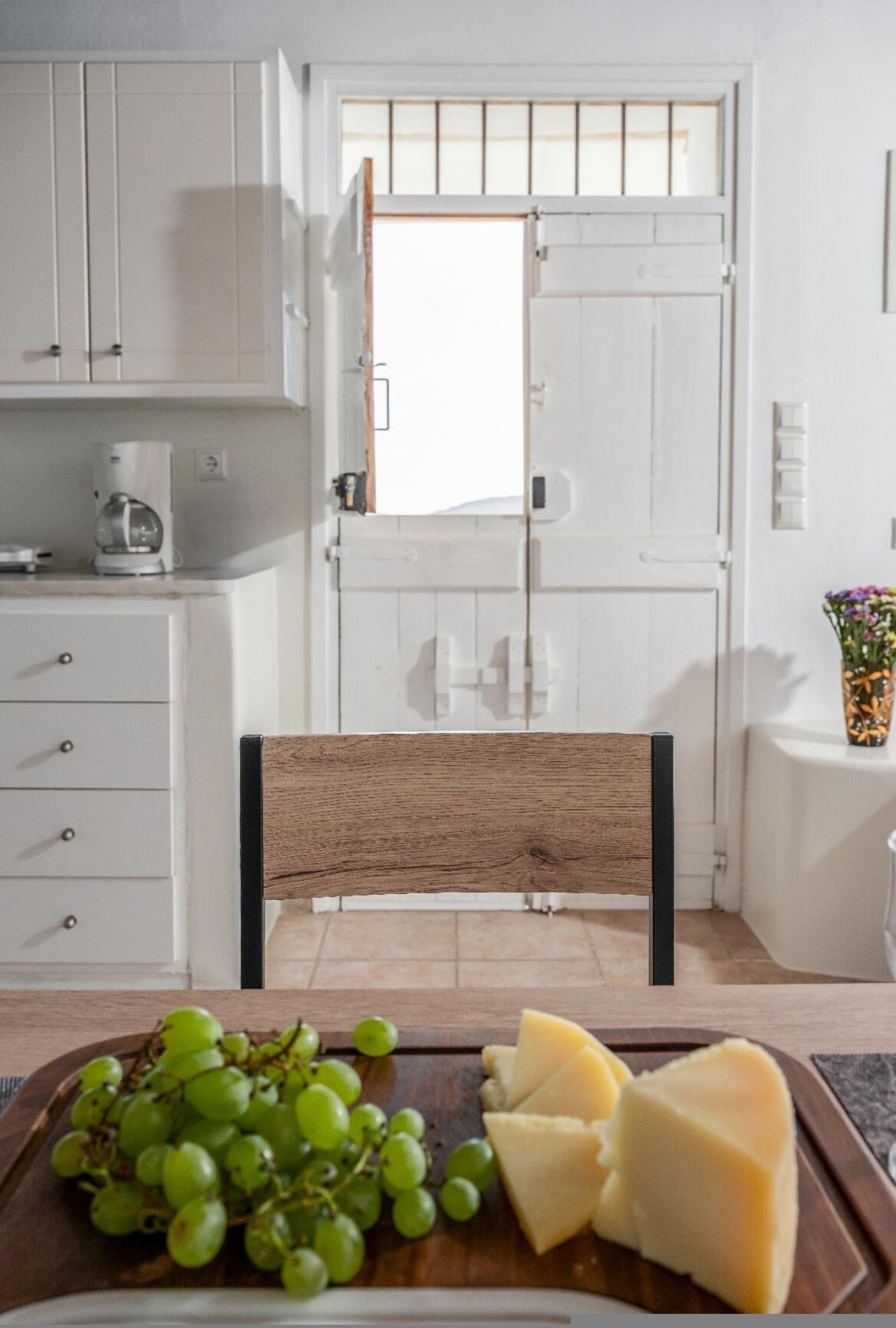 A kitchen with white cabinets, a wooden chair, a cutting board with cheese and grapes, and a coffee maker.