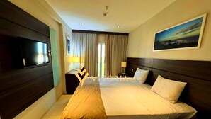 Habitación de hotel con una cama grande, un televisor de pantalla plana montado en la pared, un escritorio con lámpara y una ventana con cortinas.