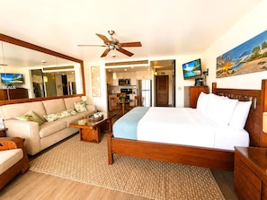 A hotel room with a bed, a sofa, a coffee table, a TV, a ceiling fan, and a kitchen area visible through an open door.