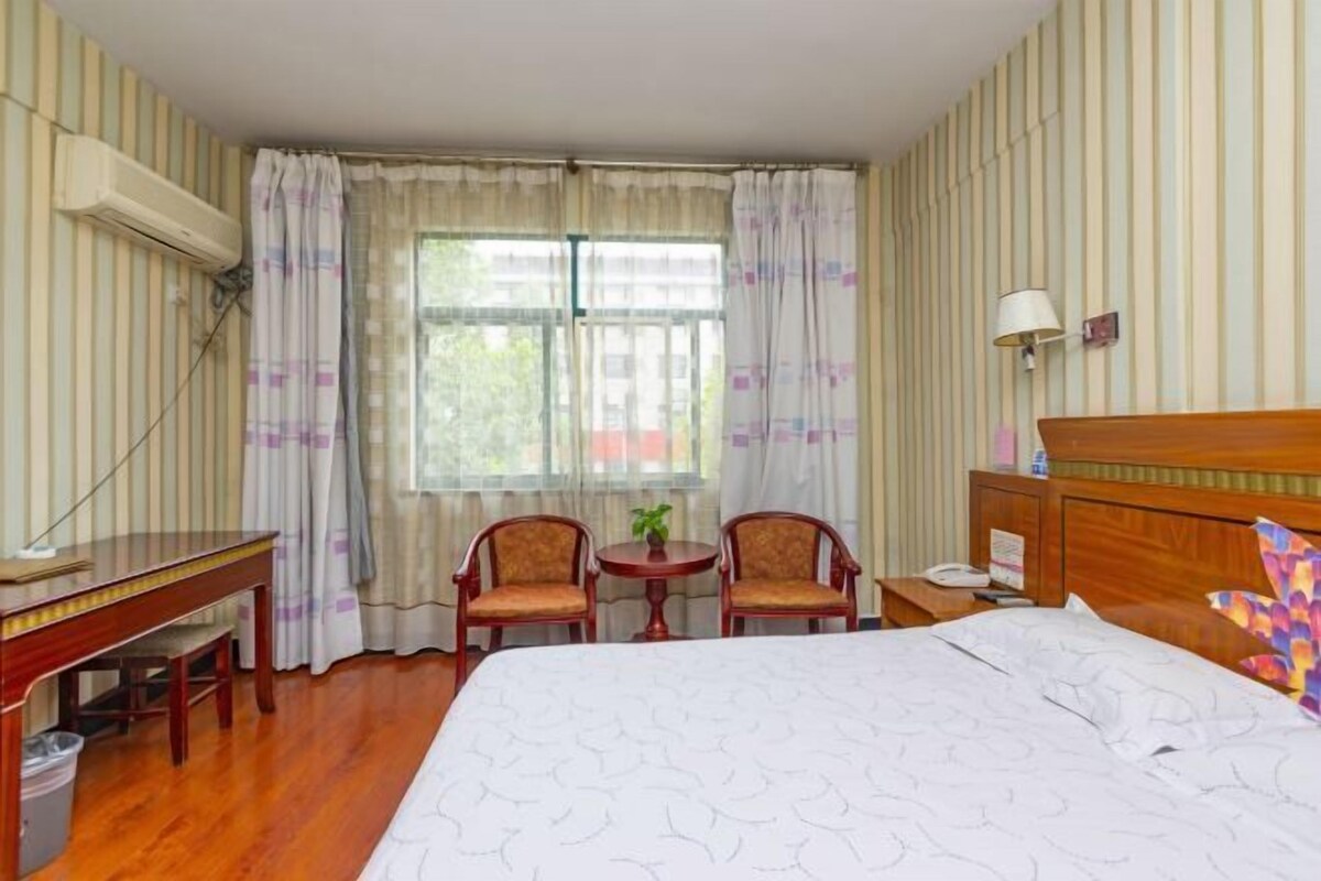 Une chambre d’hôtel avec un lit, un bureau, deux chaises, une petite table avec une plante, une lampe et une fenêtre avec des rideaux.