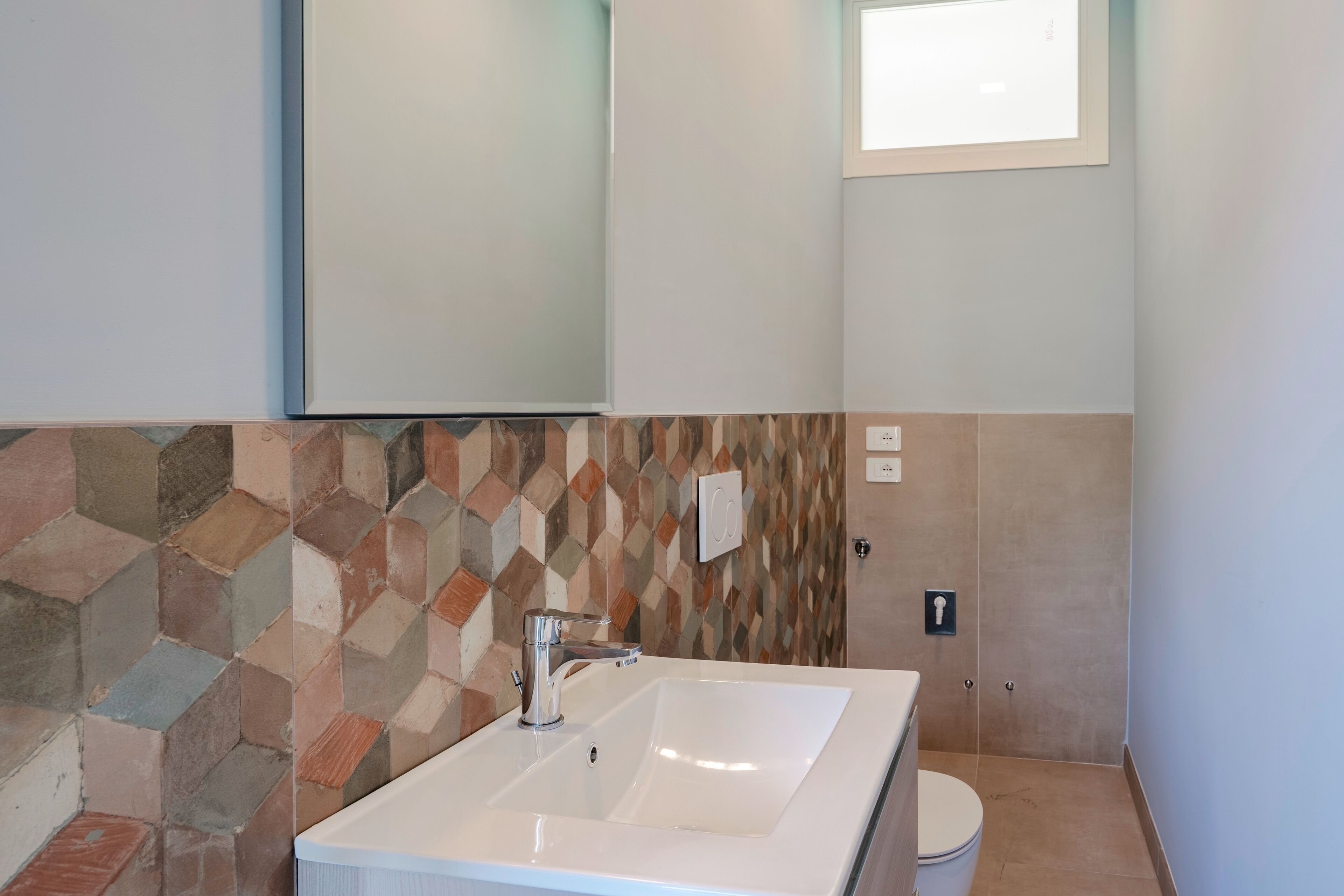 Een badkamer met een witte wasbak, een moderne kraan en een betegelde muur met een geometrisch patroon.