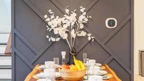 Une longue table en bois, dressée avec de l’argenterie blanche, des assiettes, des bols et des verres, un vase rempli de fleurs blanches et un panier de fruits.