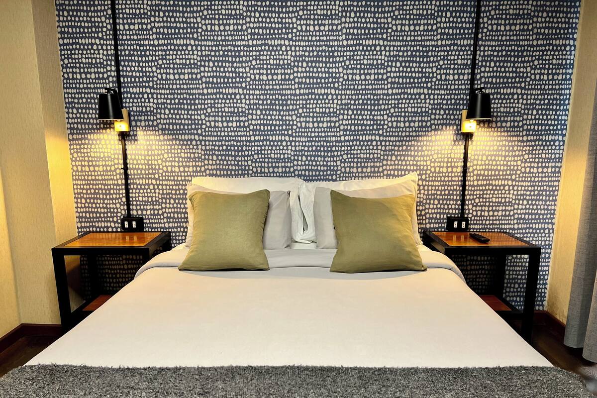 A neatly made bed with two pillows, a bedside table with a lamp, and a patterned wallpaper.