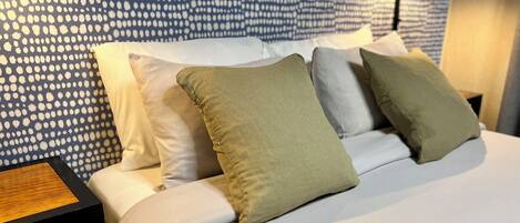 A neatly made bed with white and green pillows, a wooden nightstand, and a patterned wall.