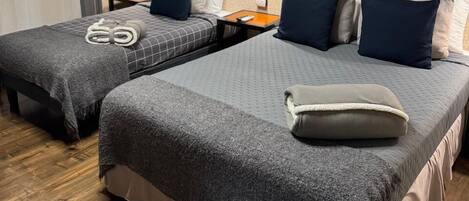 Habitación de hotel con dos camas, cada una con una manta gris y almohadas, suelo de madera y pared beige con textura.
