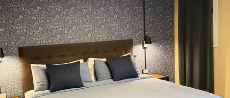 Una cama bien hecha con una manta gris y almohadas, un cabecero de madera y una lámpara de techo circular.