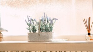 Uma janela com vidro fosco, um vaso com lavanda, um castiçal de vidro e um difusor com palitos de junco.