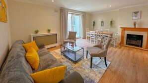 Un salon comprenant un canapé, des fauteuils, une table basse, une cheminée, un coin repas avec une table et des chaises, et une fenêtre avec des rideaux.