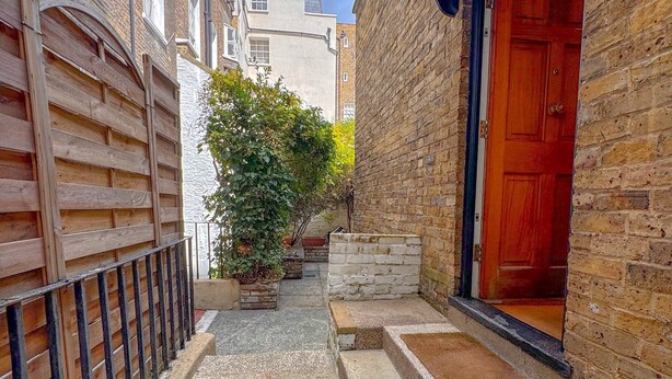 Une ruelle étroite, bordée d’une clôture en bois d’un côté et d’un bâtiment en briques de l’autre. On y aperçoit un petit jardin avec des plantes en pot et une porte rouge.