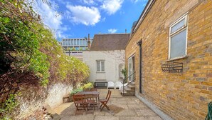 Une cour avec une terrasse en pierre, une table en bois et des chaises. Un mur de briques se trouve d’un côté, et un bâtiment blanc se dresse à l’arrière-plan.