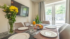 A dining area with a table set for four, a vase of yellow flowers, a bottle of orange juice, and a bowl of fruit. - Charming Modern Retreat with Private Garden - Pass the Keys (Market Drayton)