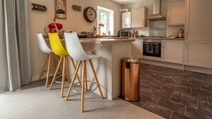 A modern kitchen with a central island, white chairs, a red lamp, and a copper trash bin. - Charming Modern Retreat with Private Garden - Pass the Keys (Market Drayton)