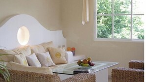 A room with a white sofa, wicker chairs, a glass table with fruit, and a window with a view of greenery. - Presidential Junior Suite - Cabarete (Puerto Plata)