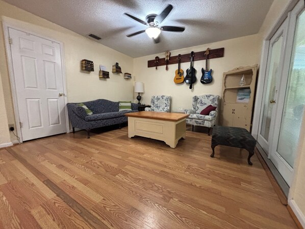 A living room with a wooden floor, a sofa, a coffee table, a chair, a ceiling fan, and a wall-mounted guitar rack.