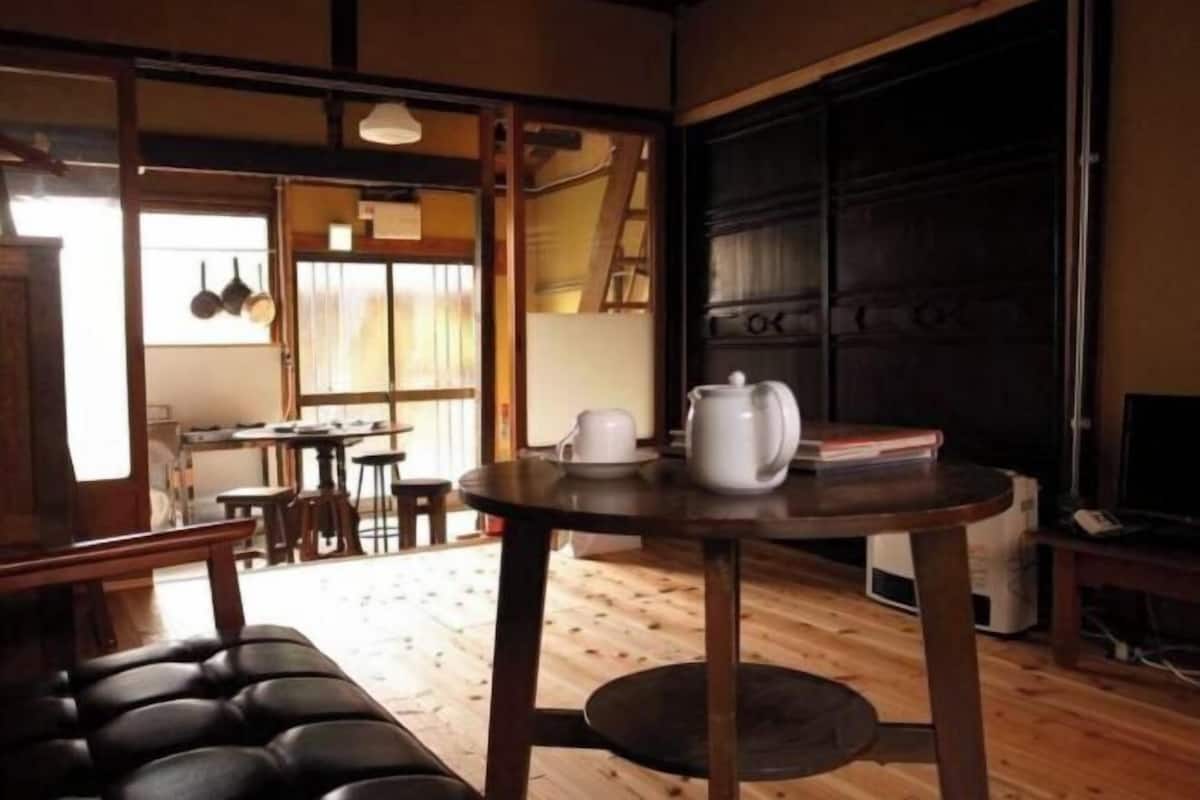 A room with a wooden floor, a round table with a white pitcher and cup, a cushioned bench, and a dark door.