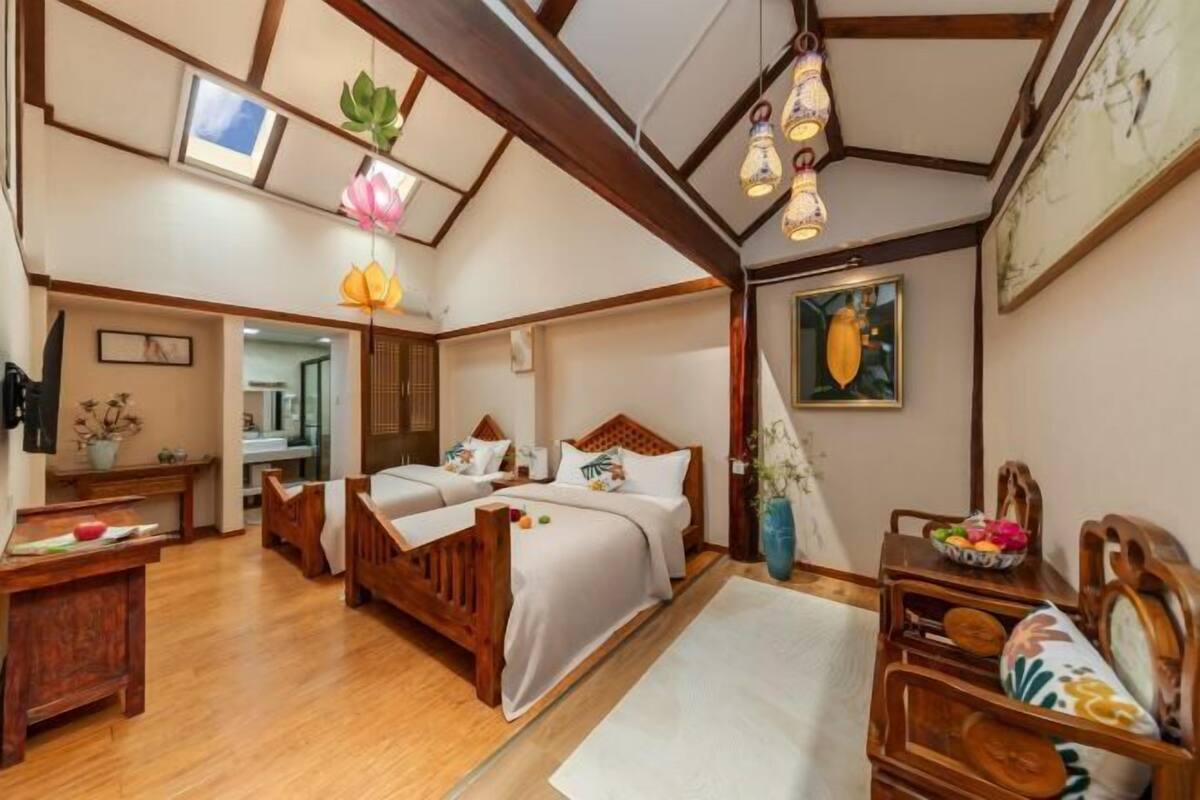 A room with wooden flooring, a bed with a headboard, a desk, a chair, a vase with flowers, and a painting on the wall.