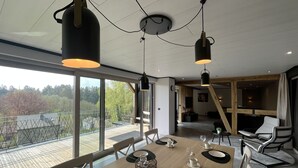 A modern dining area with a large table set for six, large windows offering a view of the outdoors, and a loft-style living area in the background. - Lu T'Chêne - dans le Village d'Opont (Opont)