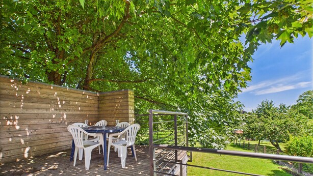 Un patio exterior con terraza de madera, una mesa con sillas y barandilla metálica que da a un área cubierta de césped.