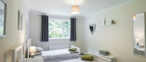 A bedroom with two beds, a window with curtains, a radiator, a bedside table with a lamp, a heart-shaped decoration, and a shelf with a plant.