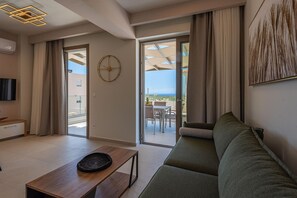 A modern living room with a sofa, a coffee table, and a dining area visible through a glass door. - Athel Panoramic View Villas Athina (Rethymno)