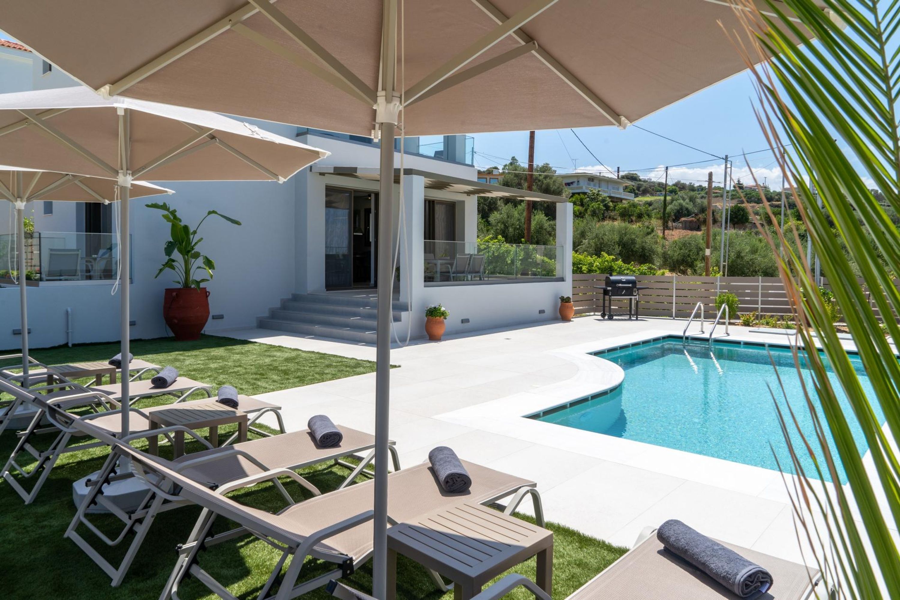 Un área de piscina moderna al aire libre con sillones de descanso y sombrillas, una piscina azul cristalina y una casa al fondo.