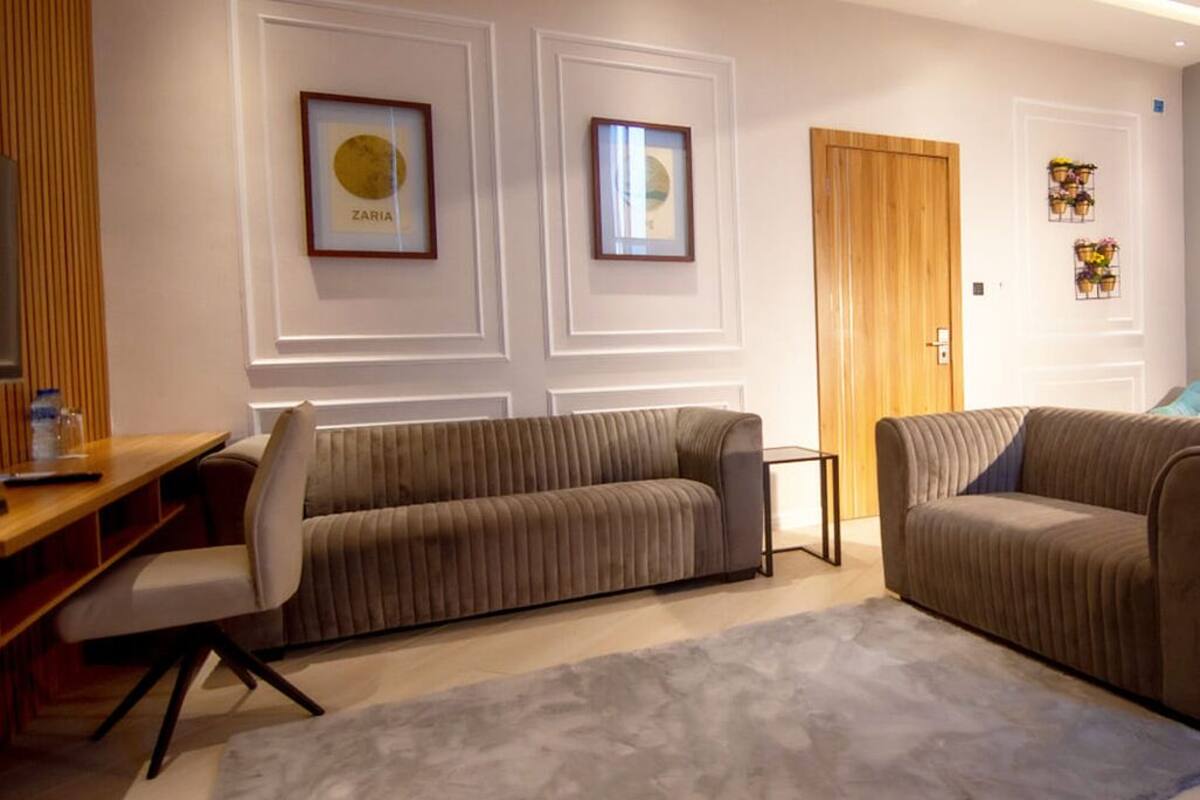 Un salon moderne avec deux canapés, une table basse et un grand tapis. Les murs sont ornés de tableaux encadrés et une porte en bois est visible.