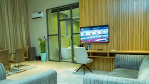 A modern living room with a television, seating, and a glass door leading to a bathroom. - 649 Suites (Asaba)