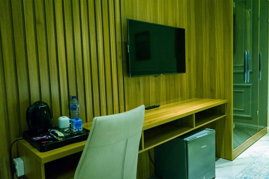 A hotel room with a wooden panel wall, a flat-screen TV mounted above a wooden desk, a small refrigerator, and a chair.