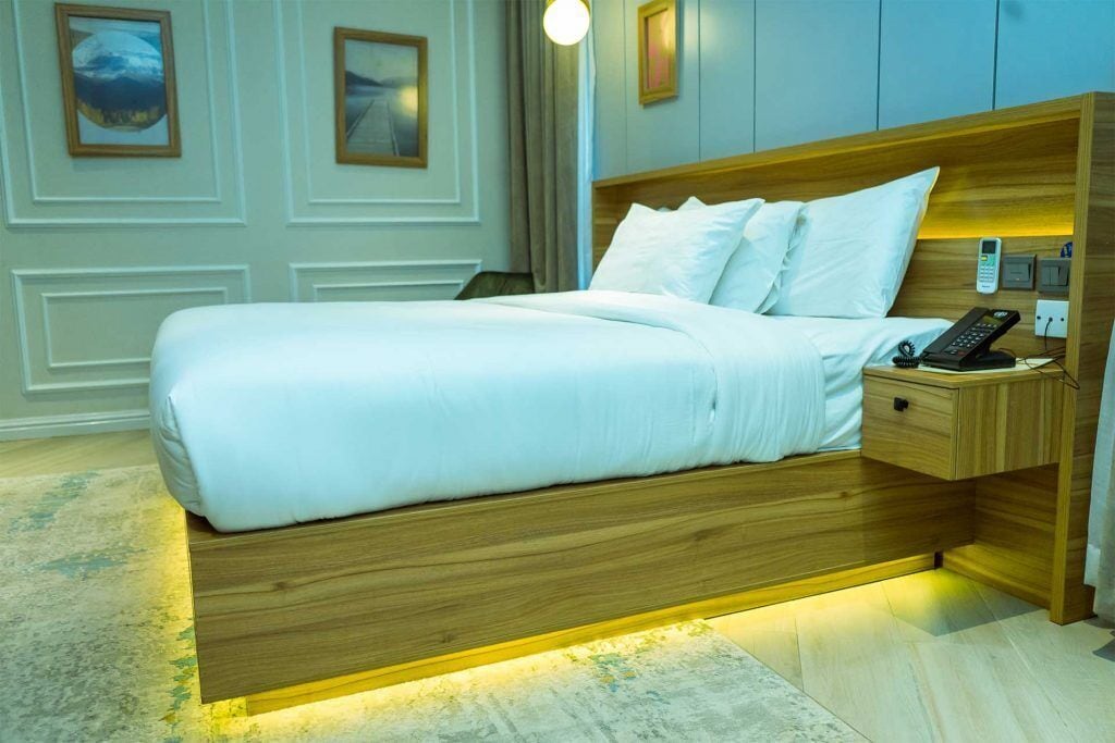 A neatly made bed with white linens, a wooden headboard, and a bedside table with a phone and a lamp.