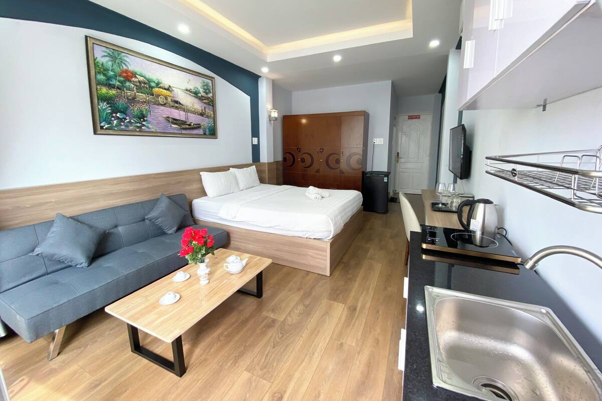Ein modernes Hotelzimmer mit einer Couch, einem Bett, einem Esstisch mit Tee-Service, einer Küchenzeile mit Spüle und einem Gemälde an der Wand.