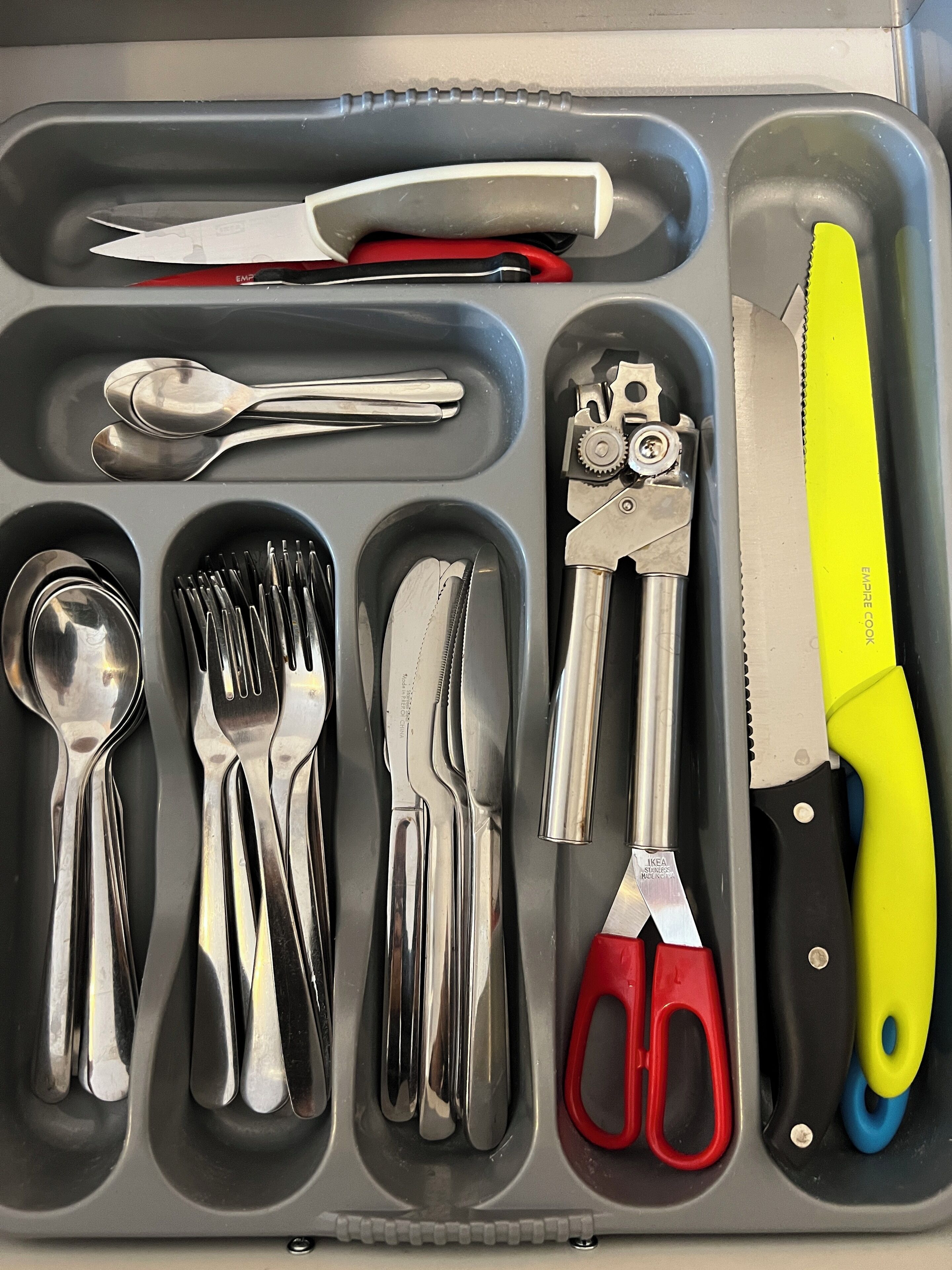 A drawer containing various utensils and tools including spoons, forks, knives, scissors, and a can opener.