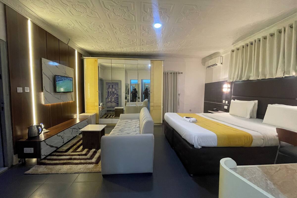A modern hotel room with a large bed, a sofa, a TV, and a bathroom visible through a glass door.