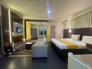 A modern hotel room with a large bed, a sofa, a TV, and a bathroom visible through a glass door. - Flora Suites (Lagos)