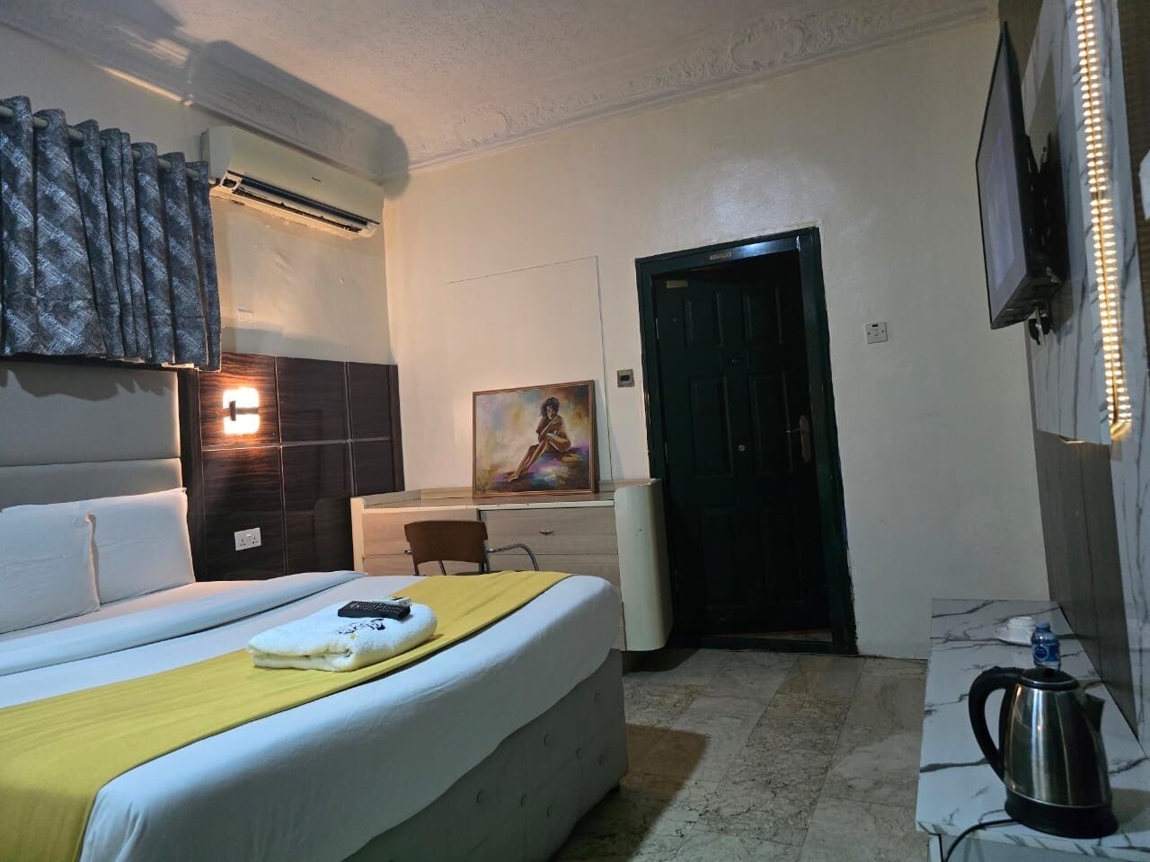 A hotel room with a bed, a desk, a chair, an air conditioner, a painting, a door, and a kettle.