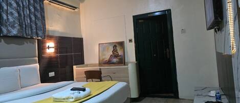 A hotel room with a bed, a desk, a chair, an air conditioner, a painting, a door, and a kettle.