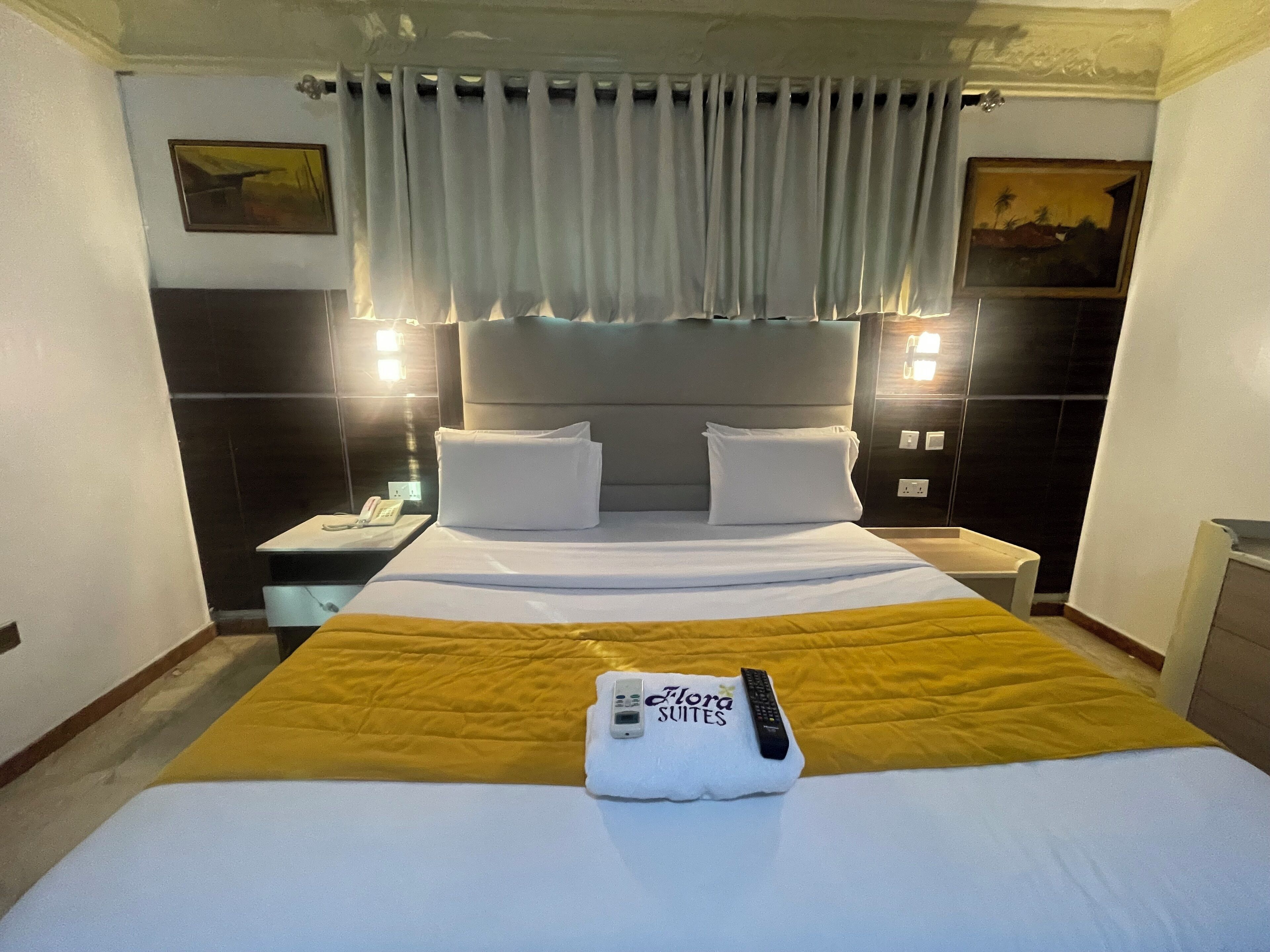 A hotel room with a bed, a nightstand, a telephone, a remote control, and a painting on the wall.