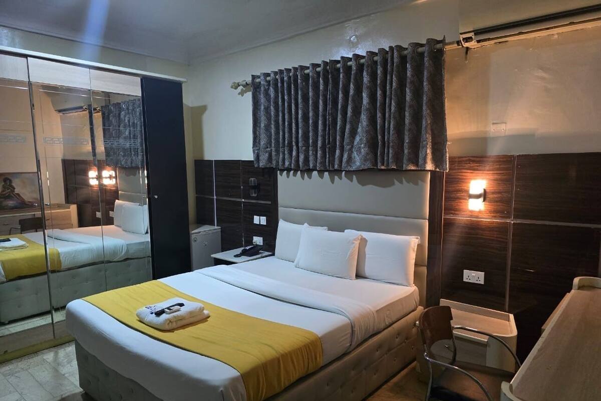 A hotel room with a bed, bedside table, mirror, and a wall-mounted light fixture.