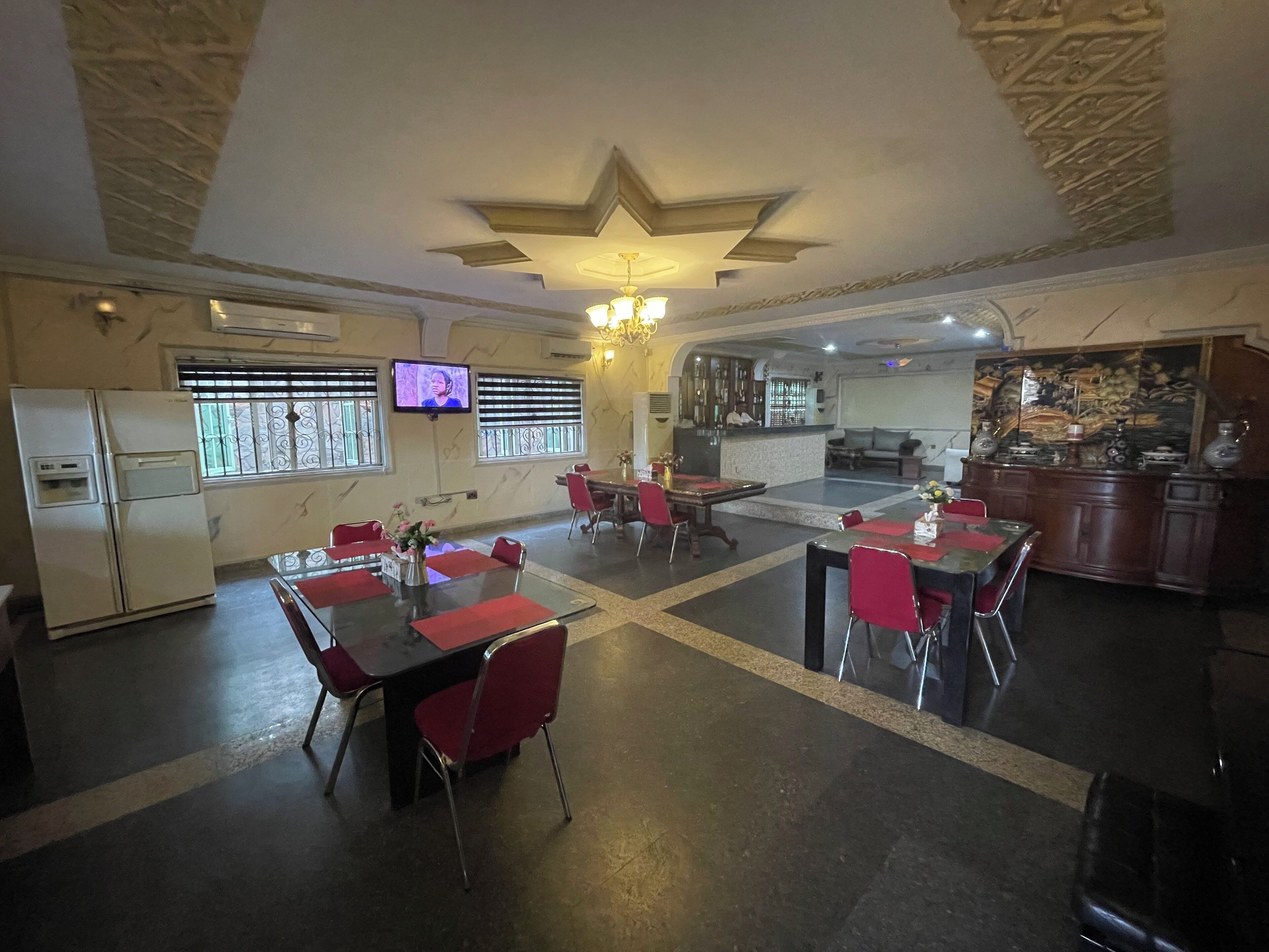 A restaurant with multiple dining tables and chairs, a television mounted on the wall, and a kitchen area visible in the background.