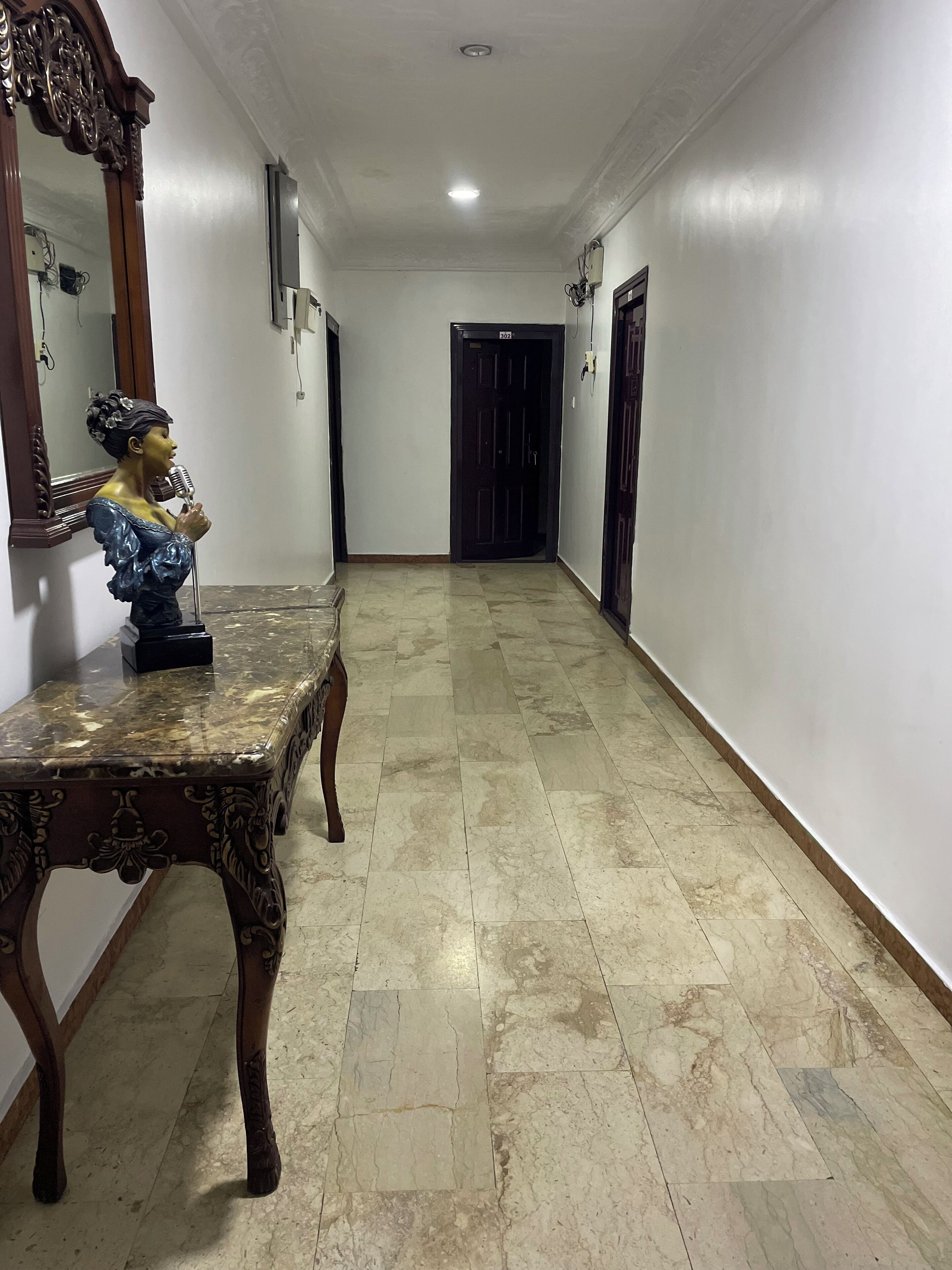 Un pasillo con suelo de mármol, una mesa de madera con una escultura de busto, un espejo y una puerta al final.