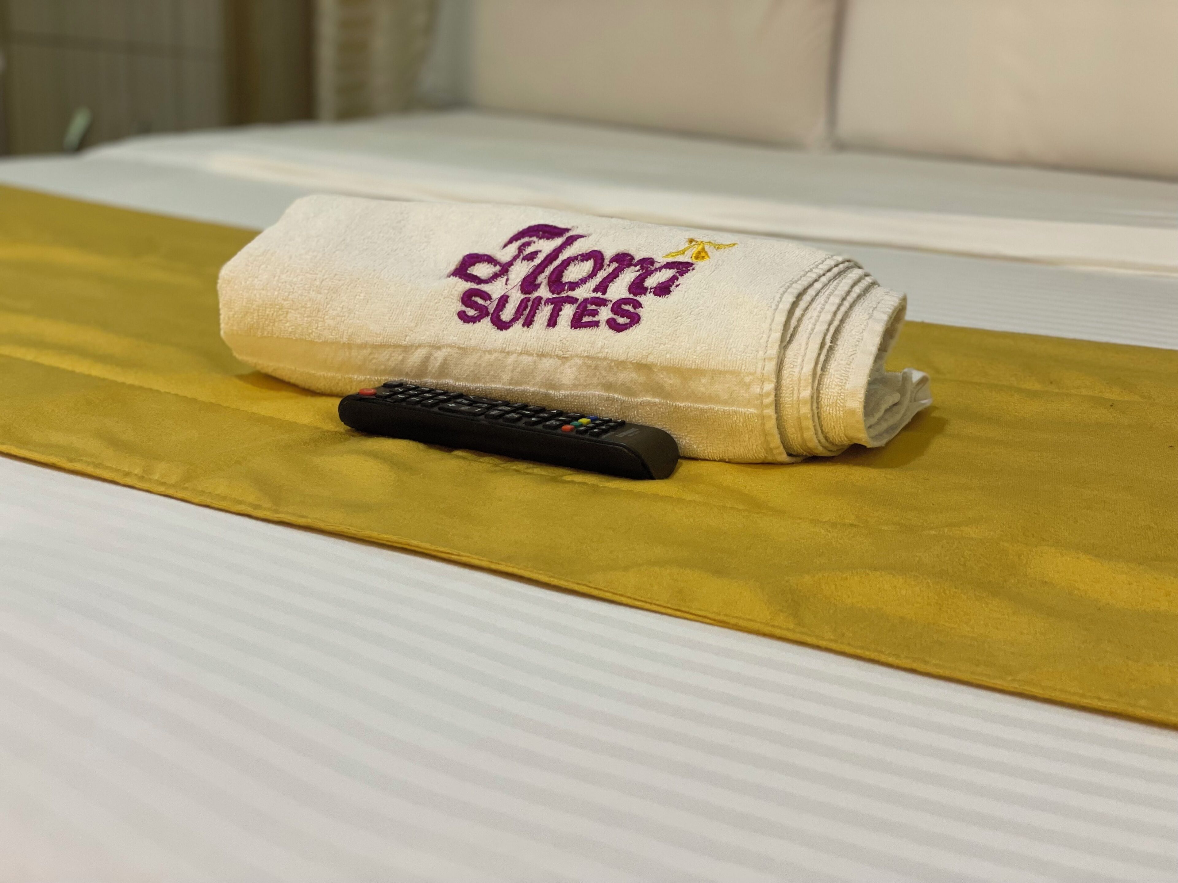 A rolled towel with "Flora Suites" embroidered on it, placed on a bed with a gold-colored bed runner.