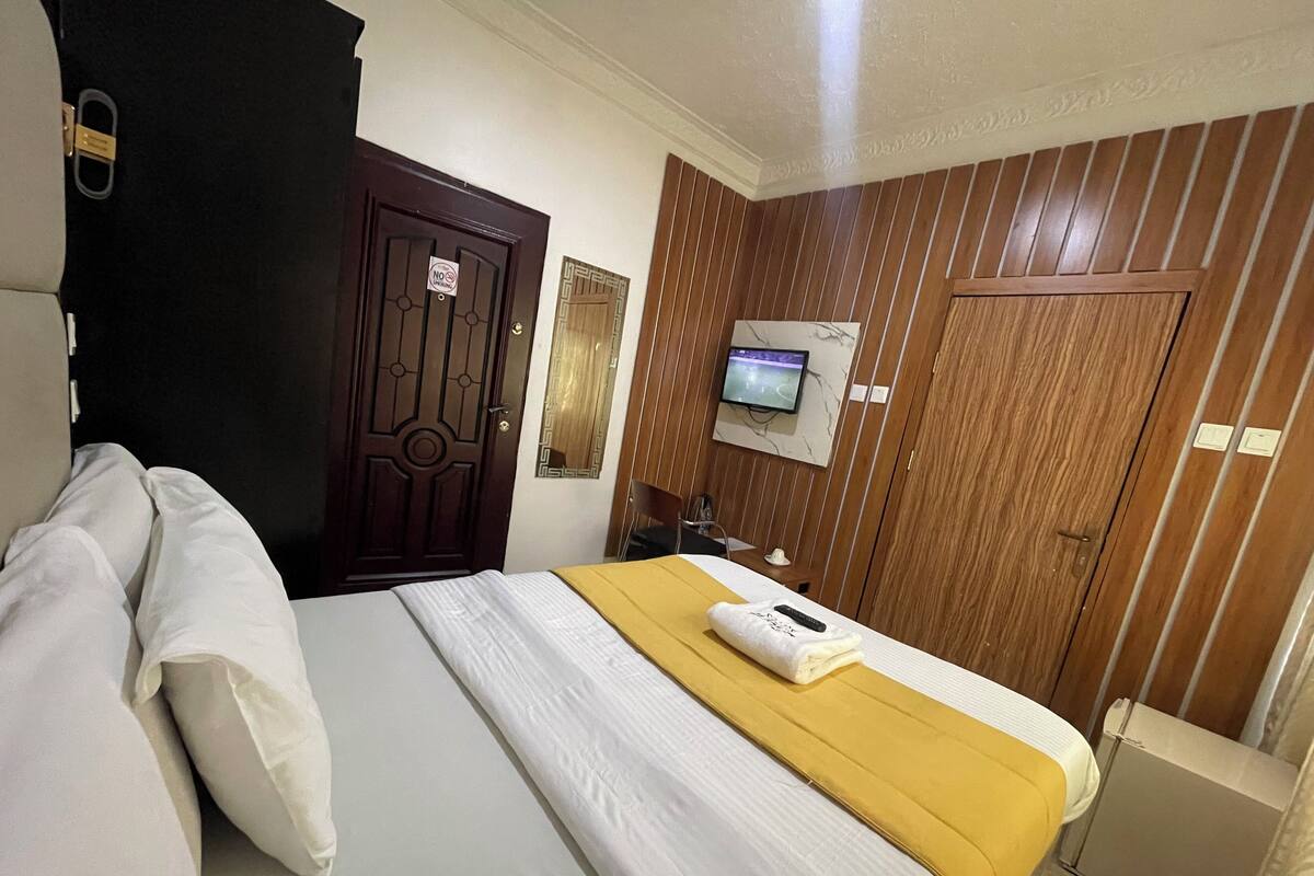 A hotel room with a bed, a TV mounted on the wall, a wooden wardrobe, and a door with a 'No Entry' sign.