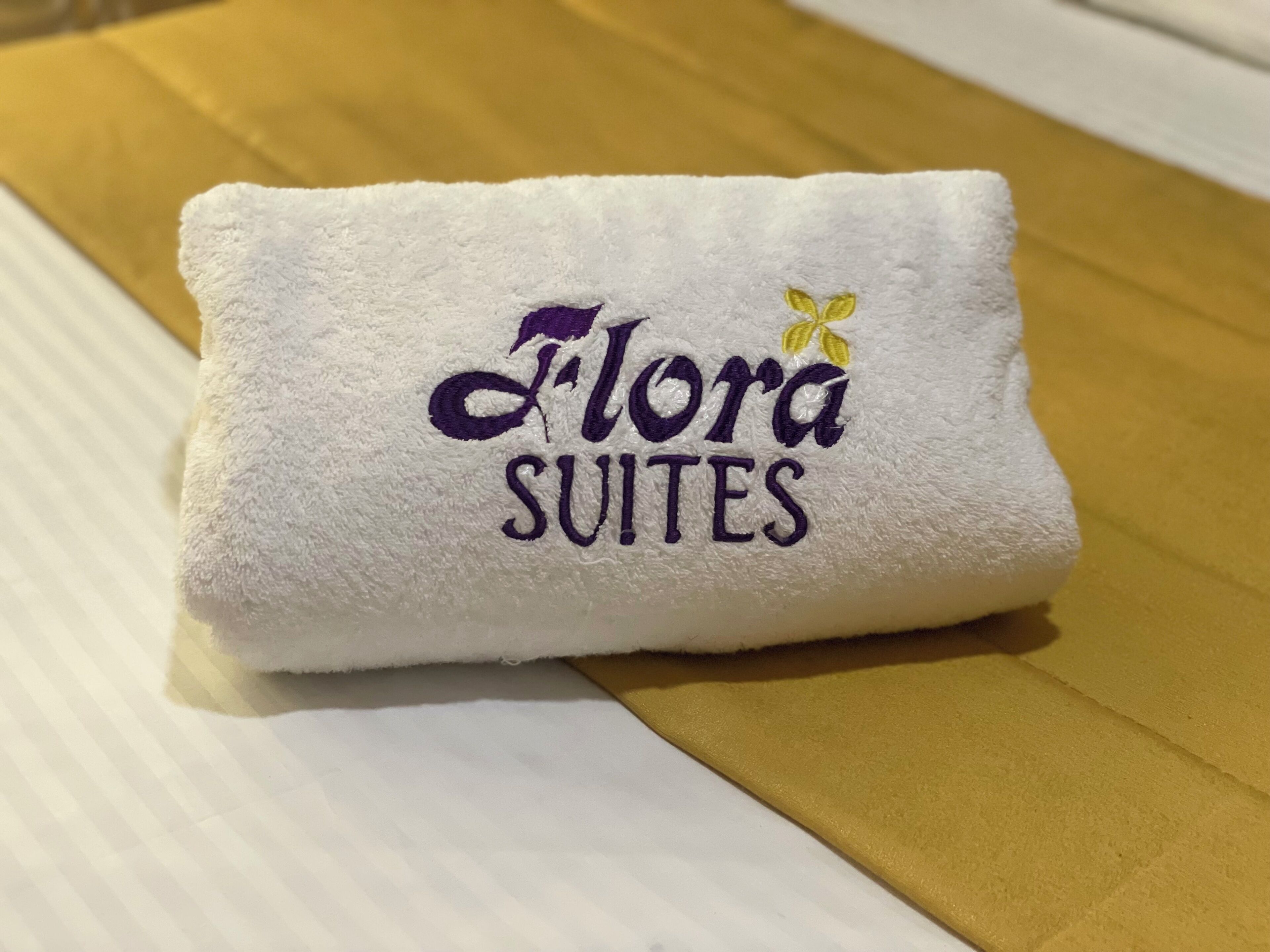 A folded white towel with embroidered text reading "Flora Suites" and a small yellow flower.
