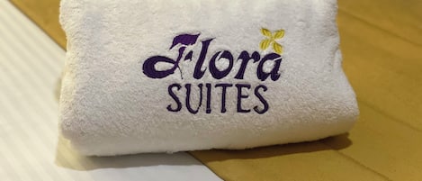 A folded white towel with embroidered text reading "Flora Suites" and a small yellow flower.