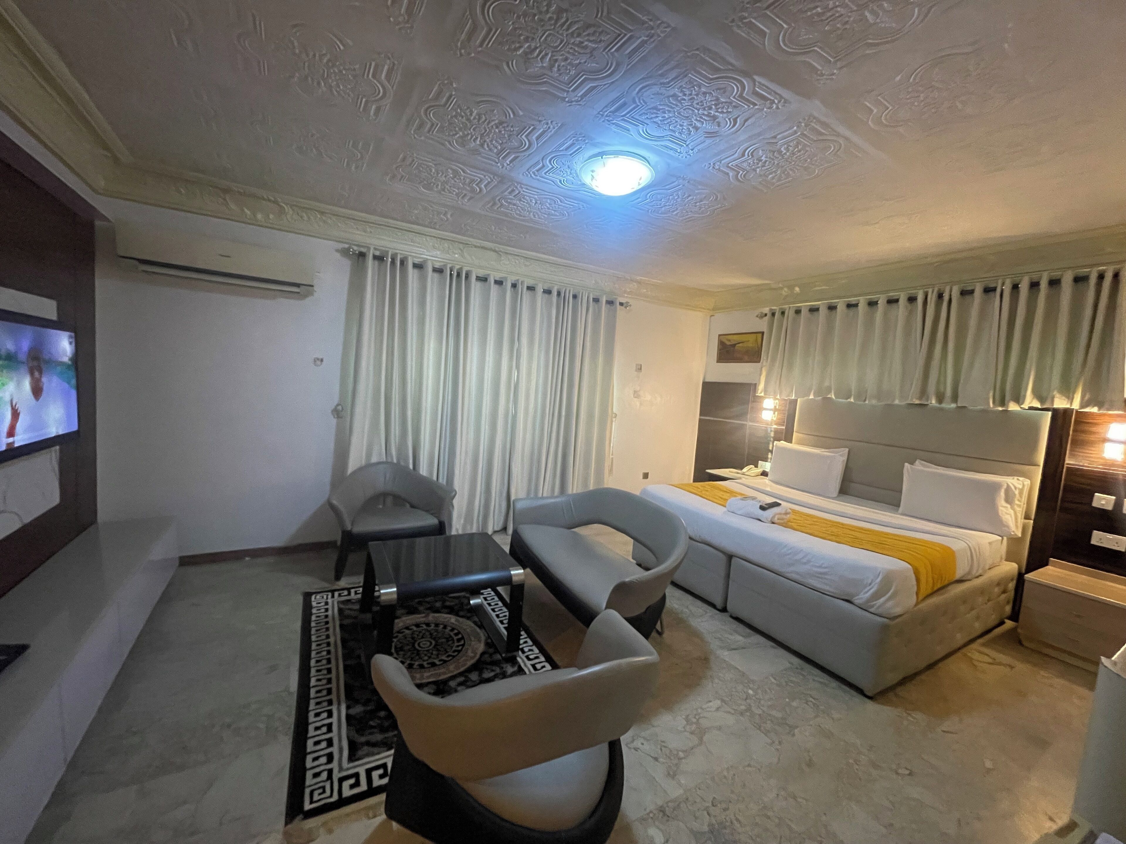 Een hotelkamer met een bed, een bureau, twee stoelen, een televisie en een plafond met gedetailleerde patronen.